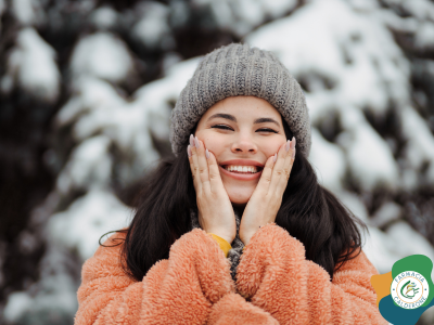 Donna sorridente con cappello di lana e giacca calda che si tocca il viso all’aperto, simbolo della cura della pelle contro il freddo.